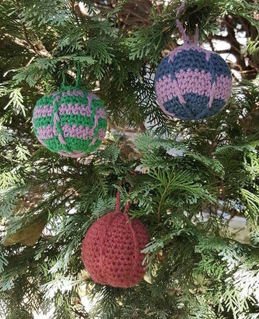 Drei gehäkelte Weihnachtskugeln (grün-rosa, blau-rosa, rot) an einem Nadelzweig