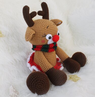 Sitzendes Amigurumi-Rentier mit roter Nase und rot-grünem Schal auf weißem Kunstfell