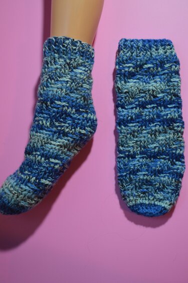 Nahaufnahme zweier gehäkelter Socken in Blautönen, einer am Fußmodell, einer flach liegend