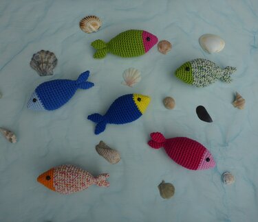 Sieben gehäkelte Amigurumi-Fische in verschiedenen Farben mit Muscheln auf hellem Stoff