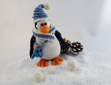 Gehäkelter Pinguin mit Mütze, Schal und kleinem blauen Vogel in einer Winterlandschaft mit Zapfen und gehäkelten Schneebällen.