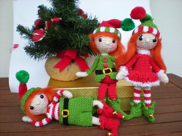 Drei festliche Amigurumi-Wichtel mit orangefarbenem Haar, rot-grünen Kleidern und Bommelmützen neben einem kleinen geschmückten Tannenbaum