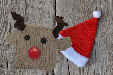 Gestrickter Rentierkopf mit Geweih und roter Knopfnase auf Holzhintergrund.