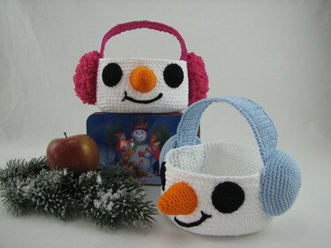 Zwei gehäkelte Schneemann-Körbchen mit pinken und blauen Ohrenschützern und Henkeln vor dezenter Weihnachtsdekoration.