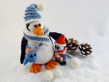 Gehäkelter Pinguin mit Bommelmütze, kleinem Vogel und blauem Kuscheltier auf künstlichem Schnee neben Tannenzapfen
