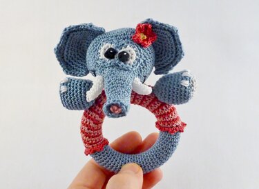 Gehäkelte Elefantenrassel, blau‑grau, mit roter Blüte und gestreiftem Greifring, in einer Hand gehalten.