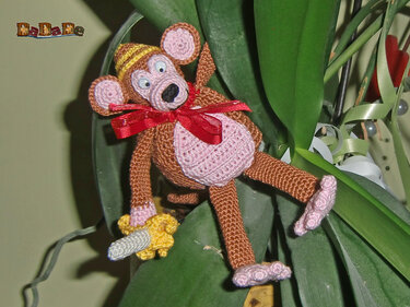 Gehäkelter brauner Amigurumi-Affe mit rosa Bauch, gelber Mütze, roter Schleife und kleiner Banane zwischen grünen Blättern