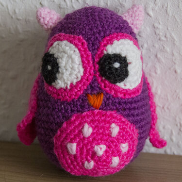 Gehäkelte Amigurumi-Eule in Lila und Pink mit großen weißen Augen, schwarzen Pupillen und orangefarbenem Schnabel auf einer Holzoberfläche vor heller Wand.