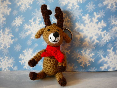 Gehäkeltes braunes Rentier-Amigurumi mit rotem Schal als Schlüsselanhänger vor Schneeflocken-Hintergrund