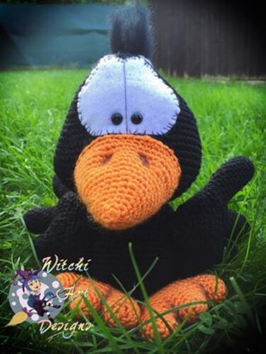 Gehäkelter schwarzer Rabe mit orangefarbenem Schnabel und Füßen, weißer Gesichtszeichnung und Knopfaugen sitzt im Gras.