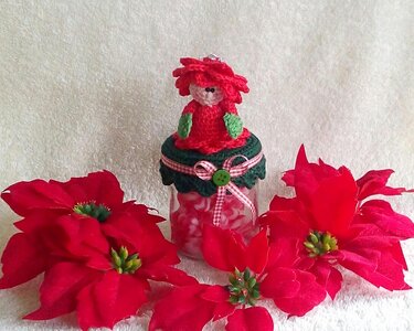 Roter gehäkelter Wichtel auf verziertem Glas, umgeben von roten Weihnachtssternen