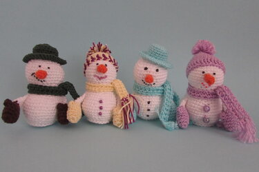 Vier gehäkelte Amigurumi‑Schneemänner mit bunten Mützen, Schals und Fäustlingen vor hellblauem Hintergrund.