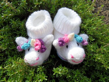 Weiße gehäkelte Babyschuhe mit Einhornköpfen und bunten Mähnen auf grünem Laub
