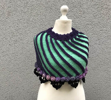 Gestreifter Poncho in Mintgrün und Violett mit gerüschtem Kragen und gehäkelter Blumenbordüre auf einer Büste.