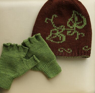 Braune gestrickte Mütze mit grünem Intarsienmuster neben grünen fingerlosen Handschuhen
