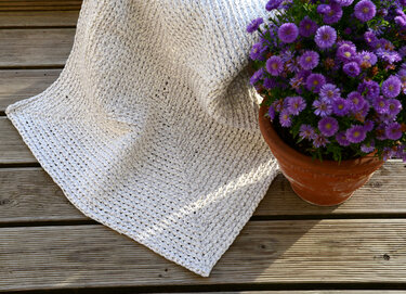 Cremefarbenes gehäkeltes Plaid auf Holzterrasse neben Terrakottopf mit violetten Blumen