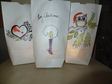 Drei beleuchtete weiße Papierbeutel mit handgemalten Weihnachtsmotiven: Herz, Kerze, Eule mit Weihnachtsmütze.