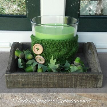 Grüne Kerze im Glas mit gestrickter Manschette und Holzknöpfen, umgeben von Eicheln und Eichenblättern auf einem Holztablett.