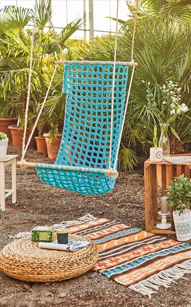 Detailreicher gehäkelter Hängesessel in Türkis mit Seilaufhängung und Holzstangen vor üppigen Pflanzen.