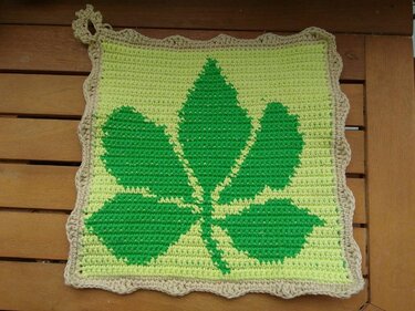 Quadratischer gehäkelter Topflappen mit grünem Blatt auf hellgrünem Grund und beigem Wellenrand, mit Schlaufe, auf Holzoberfläche.