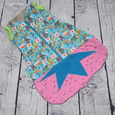 Babyschlafsack aus hellblauem Koala-Stoff mit rosa Unterteil, großer Stern-Applikation und Reißverschluss auf Holzuntergrund.