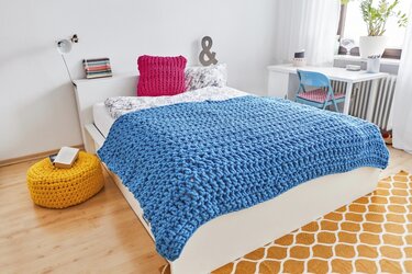 Grob gehäkelte blaue Decke auf weißem Bett mit pinkem Kissen und gelbem Pouf