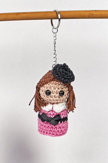 Gehäkelter Amigurumi-Puppen-Schlüsselanhänger mit rosa Kleid, braunen Zöpfen, weißem Kragen und schwarzer Baskenmütze an Metallring und Kette