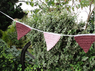 Gehäkelte Wimpelkette mit rosa und bordeauxfarbenen Dreiecken an weißer Kordel vor einem grünen Strauch.