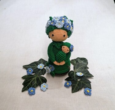 Gehäkelte Amigurumi-Puppe im grünen Outfit mit blau-weißem Blumenkranz, die ein kleines Baby im Arm hält, umgeben von Efeublättern und Häkelblüten.