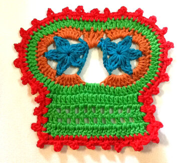 Nahaufnahme einer bunten, handgehäkelten Applikation in Zuckerschädel‑Form mit Blumenaugen und gezacktem roten Rand