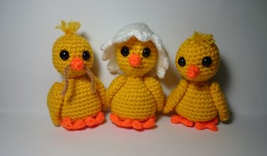 Drei gehäkelte gelbe Küken mit orangefarbenen Schnäbeln und Füßen. Das linke Küken hat braune Schnüre, das mittlere Küken trägt ein weißes Eierschalen-Häubchen.