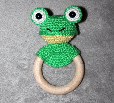 Gehäkelter grüner Frosch an einem Holzring als Beißring auf grauem Stoff