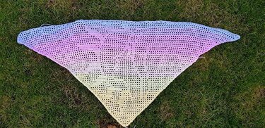 Gehäkeltes dreieckiges Tuch in Pastellfarben (Blau, Rosa, Gelb) auf Gras.