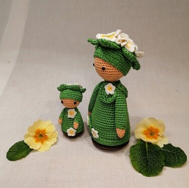 Zwei gehäkelte Amigurumi-Puppen in Grün mit weißen Blütenapplikationen und gelben Blüten daneben