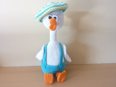 Weißes gehäkeltes Gänse-Amigurumi mit blauer Latzhose, gestreiftem Hut und orangefarbenem Schnabel und Füßen.