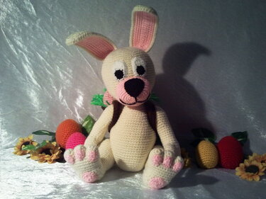 Gehäkelter Amigurumi-Hase in Creme mit rosa Pfoten und großen Ohren, sitzt vor gehäkelten Ostereiern und Sonnenblumen