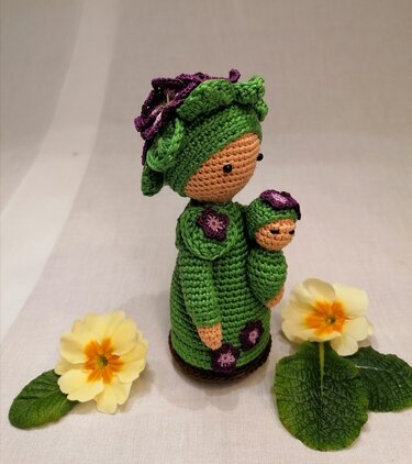 Gehäkelte Amigurumi-Puppe im grünen Kleid mit lila Blüten hält eine kleine Puppe im Arm; davor liegen zwei gelbe Blüten