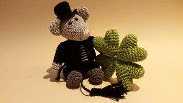 Gehäkelte Amigurumi‑Maus mit Zylinder neben einem grünen vierblättrigen Kleeblatt.