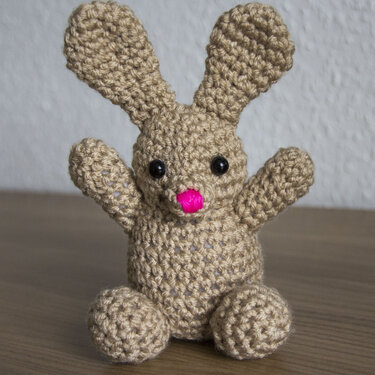 Beiger gehäkelter Amigurumi-Hase mit langen Ohren, pinker Nase und dunklen Augen, sitzend