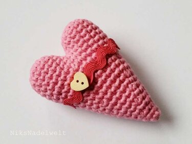 Rosa gehäkeltes Herz-Amigurumi mit rotem Zickzackband und herzförmigem Holzknopf