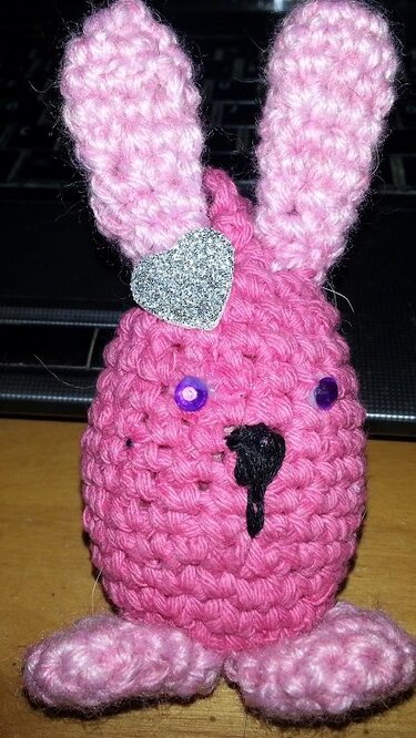 kleines rosa Amigurumi-Häschen mit sichtbaren Maschen, violetten Knopfaugen und glitzerndem Herz am Ohr