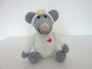 Gehäkelte graue Amigurumi-Maus im weißen Doktor-Outfit mit roter Applikation und Knopf am Kopf