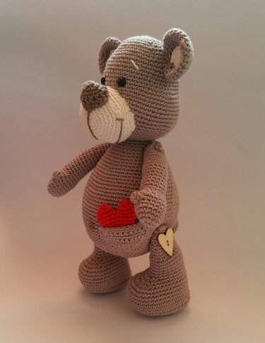 Gehäkelter Amigurumi-Teddybär in Braun mit rotem Herz in der Bauchtasche und Holz-Herzknopf am Bein