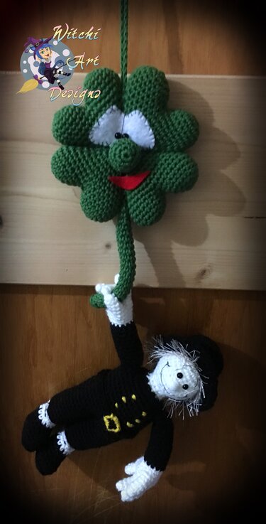 Gehäkeltes grünes vierblättriges Kleeblatt mit Augen und rotem Mund, an Schnur hängend, darunter eine gehäkelte Schornsteinfeger-Puppe in schwarzer Kleidung
