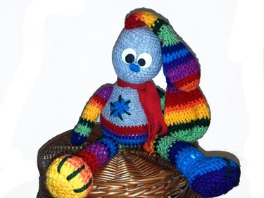 Bunter gehäkelter Amigurumi‑Hase mit langem, gestreiften Schlappohr und rotem Schal, sitzend auf einem Korb