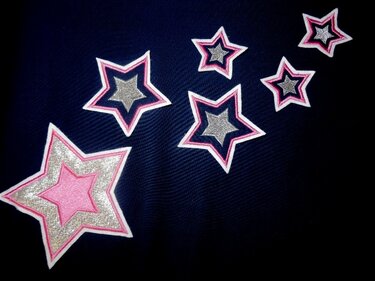 Mehrere gestickte Sterne in unterschiedlichen Größen mit pinker Mitte, silberner Füllung und weißer Umrandung auf dunkelblauem Stoff