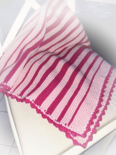 Gestrickte Babydecke in Hell- und Dunkelrosa mit diagonalen Streifen und gewellter Kante auf einem hellen Stuhl.