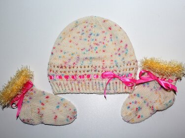 Gestrickte cremefarbene Babymütze und zwei Söckchen mit bunten Sprenkeln, rosa Satinbändern und gelbem Flauschrand