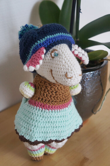 Gehäkeltes Amigurumi‑Schaf mit Bommelmütze, braunem Oberteil, hellblauem Kleid und Ringelsocken.