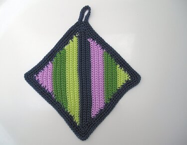 Gehäkelter rautenförmiger Topflappen mit dunkelblauer Kante, Aufhänger und diagonalen Streifen in Grün und Lila.